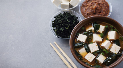Obraz na płótnie Canvas Bowl with miso soup, wakame seaweed, miso pasta, tofu and chopsticks on a gray background.