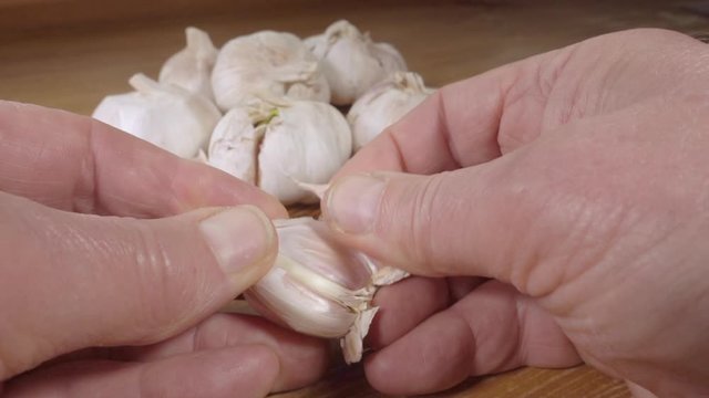 Normal speed then slow motion: Close POV shot of a man&rsquo;s hands pushing a garlic press to one side then separating cloves from a head of garlic, on a pine kitchen worktop.