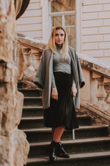 Stylish autumn fashionable girl image. Gray blazer, pleated skirt and gray coat. Blond woman/Old city background.