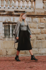 Stylish autumn fashionable girl image. Gray blazer, pleated skirt and gray coat. Blond woman/Old city background.