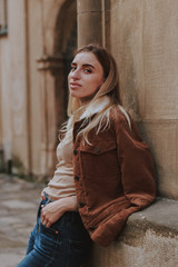 Stylish autumn fashionable image of a girl. Brown jacket with fur, jeans, boots, basic beige golf. Blonde on old city background