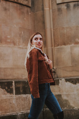 Stylish autumn fashionable image of a girl. Brown jacket with fur, jeans, boots, basic beige golf. Blonde on old city background