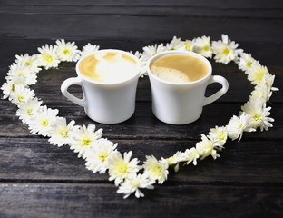 On a black background, a heart of fresh white chrysanthemum flowers and two cups of fresh coffee.