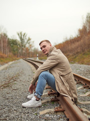 Autumn mood, young guy in a raincoat trench coat underground, street style. A young guy in a trench coat, sneakers and jeans sits on a rail of railway tracks.