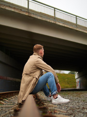 Autumn mood, young guy in a raincoat trench coat underground, street style. A young guy in a trench coat, sneakers and jeans sits on a rail of railway tracks.