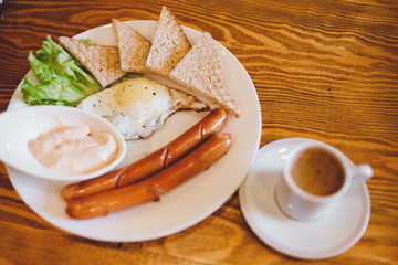 Classic English breakfast with espresso coffee in white mug, two sausages, fried eggs, toasts, lettuce on wooden background
