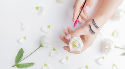 Obraz na płótnie Canvas Hands girl master eyelash extension on white flowers background with pink brush, top view. Beauty concept