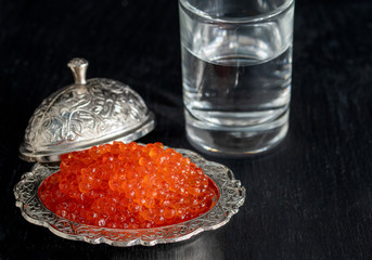 Red caviar in silver bowl with Russian vodka on black background