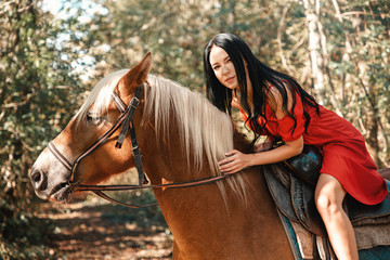 A young beautiful brunette woman in a red dress sits astride a brown horse. Summer