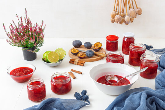 Autumn and fall seasonal kitchen work, plum compote preserving, plum jam production. Porcelain bowl and jars with compote, plums, limes, cinnamon, knife and poppies. All on white wooden background.