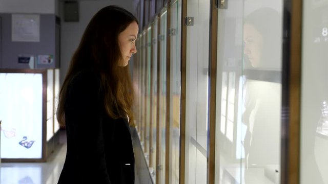 Woman stay against glass shopcase, look to jewelry assortment at airport store. Business attire of young adult lady, common shop at international Chinese terminal