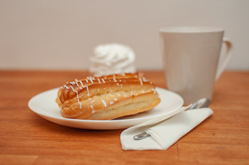 Eclair with white icing on a white plate, a teaspoon, in the background a cake and a cup of tea