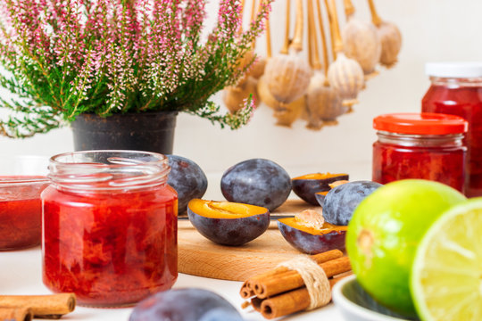 Autumn and fall seasonal kitchen work, making plum compote and jam. Porcelain bowl and jars with compote, plums, limes, cinnamon  knife and poppies. All on the white wooden background.