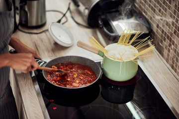 Mixed race woman in apron standing next to stove and stirring tomato sauce. On stove are saucepan and pot with spaghetti.
