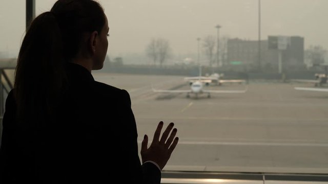 Thoughtful woman stay at window, with arm on glass. Passenger looking out to small bizjet plane parked at apron of large international airport. Silhouetted portrait shot from back