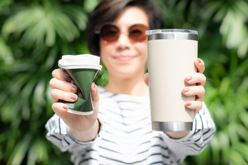 A stylish beautiful woman holding takeaway coffee cup in both hands, one is a single use paper cup with plastic lid the other one is a reusable stainless tumbler. No straw and Zero waste concept.