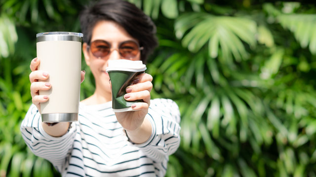 A stylish beautiful woman holding takeaway coffee cup in both hands, one is a single use paper cup with plastic lid the other one is a reusable stainless tumbler. No straw and Zero waste concept.