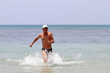 Joyful man in a swimming trunks runs into the sea in splashes of blue water, water drops on tanned skin. Paradise beach vacation, concept of swimming, happiness and freedom