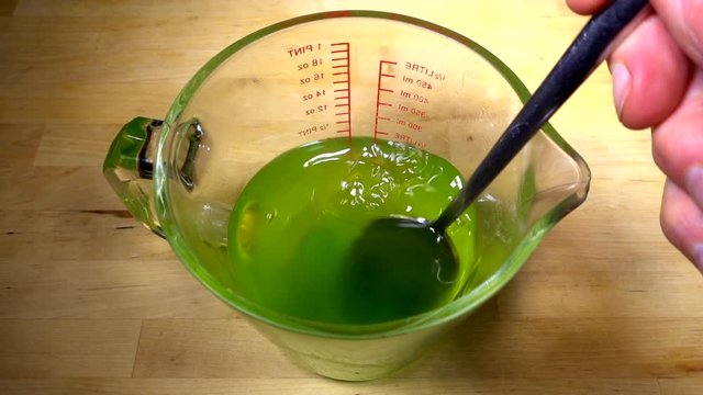 Slow motion close POV shot of a man&rsquo;s hand stirring lime green fruit jello / jelly cubes in boiling water in a glass measuring jug, to dissolve the mixture.