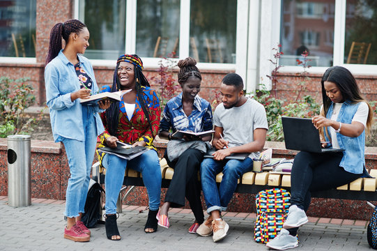 Group Of Five African College Students Spending Time Together On Campus At University Yard. Black Afro Friends Studying At Bench With School Items, Laptops Notebooks.