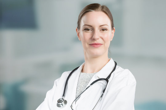 Smiling Female Family Doctor In Doctor's Overall With Stethoscope In Front Of A Clinic Room