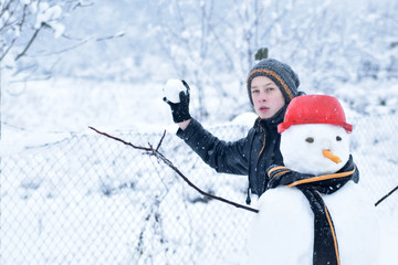 Fototapeta premium Winter teenager in a jacket and hat sculpts a snowman on a winter background with falling snow in frozen day concept