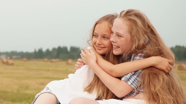 Playful girls hugging on haystack on harvesting field at village vacation. Smiling girl friends embracing on haystack background on countryside field
