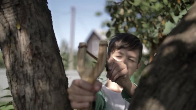 funny boy hides behind a tree and aims with a slingshot outdoors