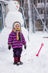 A little girl stands in a winter blizzard near a sculpture of a snowman, a child is warmly dressed in a purple jacket, overalls, hat and scarf