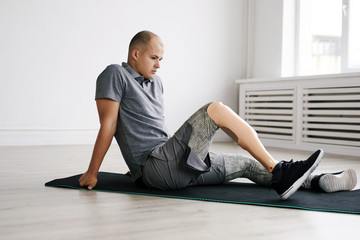 Young disabled man sitting on exercise mat and training his leg during sports training
