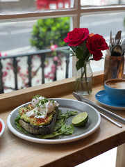Trendy breakfast layout with avotoast with poached eggs, coffee and granola on wooden windowsill against city landscape from outside.