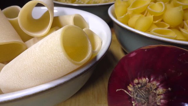 Normal speed then slow motion &ndash; Cose POV reverse dolly shot of a meal preparation, with three bowls of different types of uncooked pasta, finishing with a whole red onion.