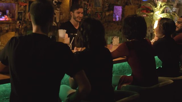Handheld shot of cheerful young bartender making cocktails and smiling while customers sitting at counter and enjoying night