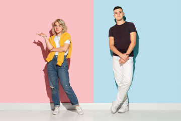 Fototapeta premium Young emotional man and woman in bright casual clothes posing on pink and blue background. Concept of human emotions, facial expession, relations, ad. Beautiful caucasian couple standing and smiling.