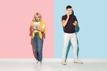Obraz premium Young emotional man and woman in bright casual clothes posing on pink and blue background. Concept of human emotions, facial expession, relations, ad. Beautiful caucasian couple using gadgets.