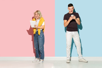Young emotional man and woman in bright casual clothes posing on pink and blue background. Concept of human emotions, facial expession, relations, ad. Beautiful caucasian couple using gadgets.