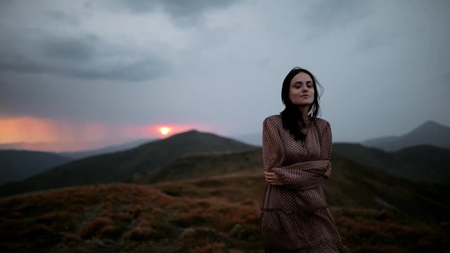 Young beautiful woman in dress walking on top of mountain during sunset. Dramatic mountain landscape with rain on the horizon. Happy and drunk on life, youth and happiness