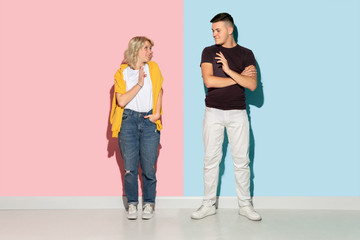 Obraz premium Young emotional man and woman in bright casual clothes posing on pink and blue background. Concept of human emotions, facial expession, relations, ad. Beautiful caucasian couple greeting and smiling.
