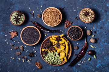 Dry whole warming Indian spices in wooden bowls on dark blue concrete background.