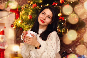 A beautiful young woman sits in a white sweater with a large cup of hot drink under the Christmas tree and dreams.