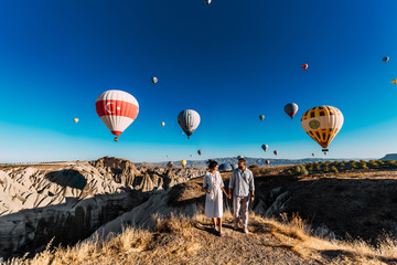 Couple in love stands on background of balloons in Cappadocia. The couple travels the world. Vacation in Turkey. Honeymoon trip. Hot air balloon flights. Happy young couple meets dawn in Cappadocia - obrazy, fototapety, plakaty