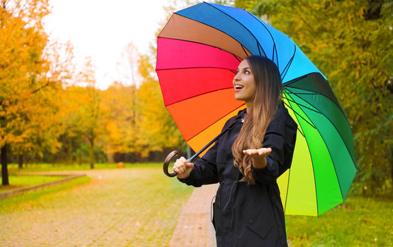 Cheerful young woman with colorful umbrella checking for rain in city park.