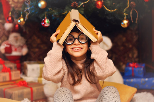 Cute little nerd girl in glasses with large lenses sits and holds an old book over her head against the background of the Christmas tree.