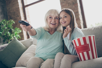 Photo of two people aged white haired granny small granddaughter sit cozy sofa watching tv eat popcorn spending summer weekend holidays together house indoors