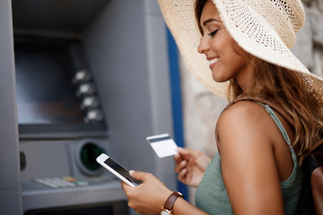 Happy woman using mobile phone while withdrawing money from ATM.