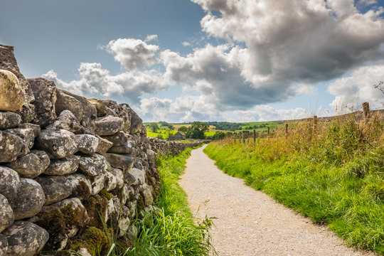 Low level view of a very old, stone wall seen extending to an abandoned farmhouse in the distance. A single track path leads the way, part of a footpath.