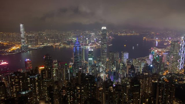Timelapse famous Hong Kong highrise buildings and towers with coloured illumination on wide harbor banks under cloudy sky at night zoom out