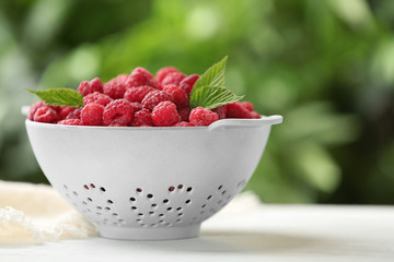 Colander with delicious ripe raspberries on white table against blurred background, space for text