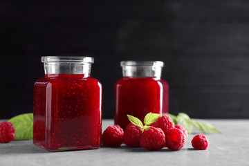 Glass jars of sweet jam with ripe raspberries and green leaves on table against black background. Space for text