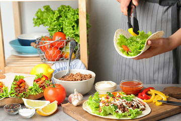 Young woman cooking delicious tortilla with meat and vegetables at table in kitchen, closeup view